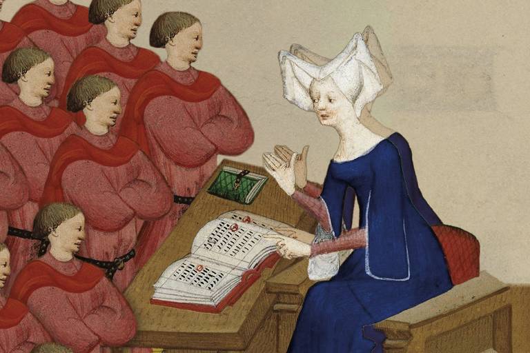 Uma mulher vestida de azul está sentada à uma mesa dando aula para outras mulheres que vestem vermelho
