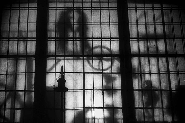 Plano do filme; sombras, ao fundo, atrás de uma janela
