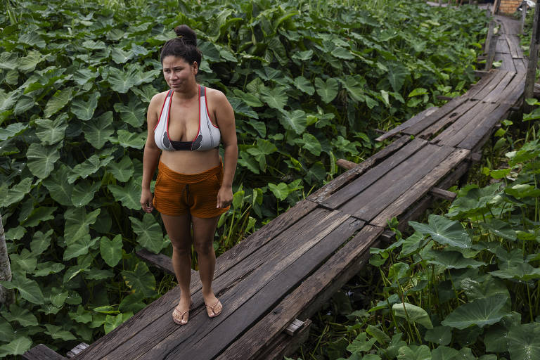 Mulher posa para foto em pé em uma ponte de madeira cercada por vegetação; ela tem o semblante sério e triste