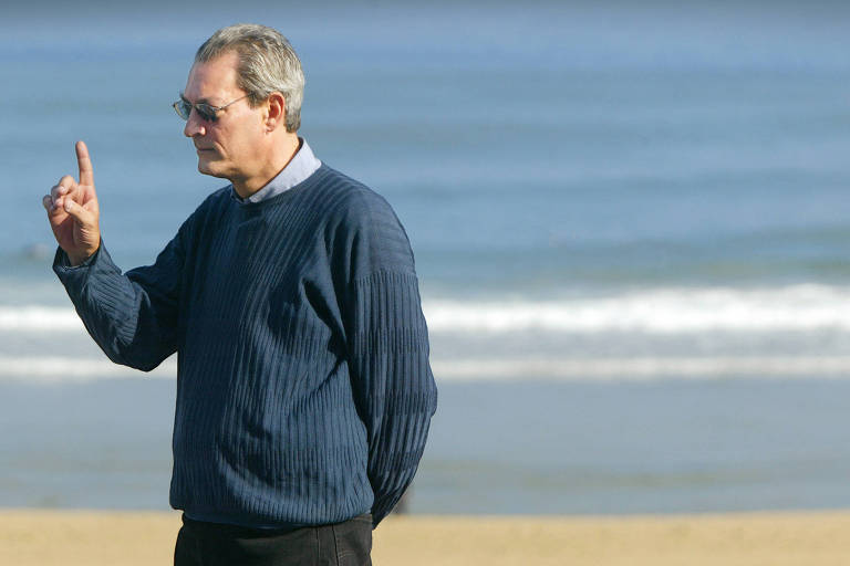 homem grisalho levanta dedo indicador da mão direita em orla de praia vestindo blusa azul e óculos escuros