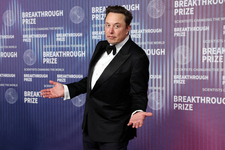 Elon Musk está vestindo smoking. Ele está com as duas mãos levantadas. Atrás dele, está um painel azul e roxo.