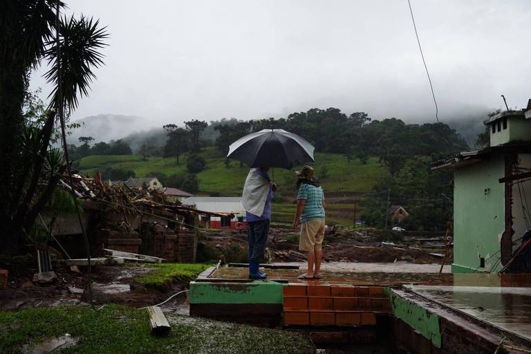 Na foto, duas pessoas estão debaixo de um guarda-chuva olhando os destroços e entulho deixados pela passagem das chuvas no Rio Grande do Sul