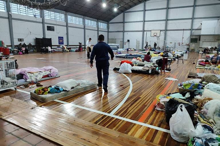 Colchões com cobertas e travesseiros espalhados no chão de um ginásio esportivo. Uma pessoa é vista de costas caminhando entre as camas improvisadas.