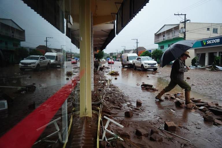 À esq., fachada de vidro de estabelecimento com calçada com destroços e chão enlameado e pessoa caminhando de guarda-chuva aberto. Ao fundo, rua de comércio e carros.