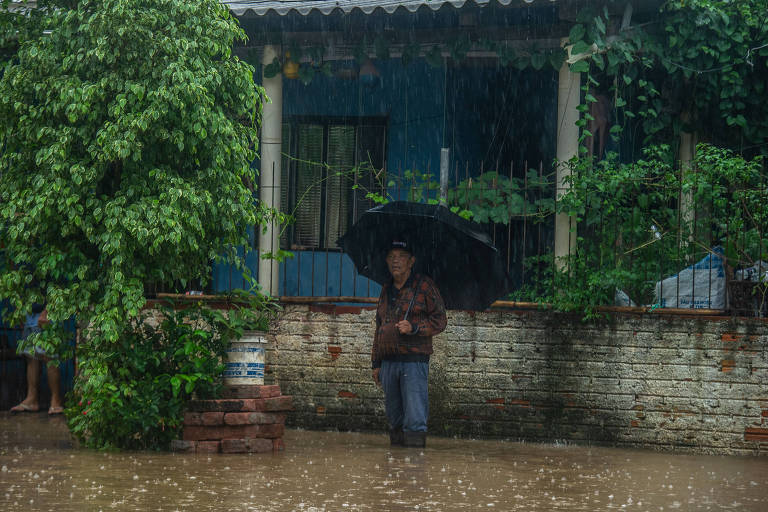 Na foto, um senhor com um guarda-chuva está na frente de uma casa com água na panturrilha