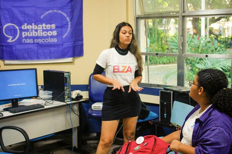 Mulher falando, posicionada no centro de uma sala de aula; no fundo, há computadores; uma menina está virada na direção dela