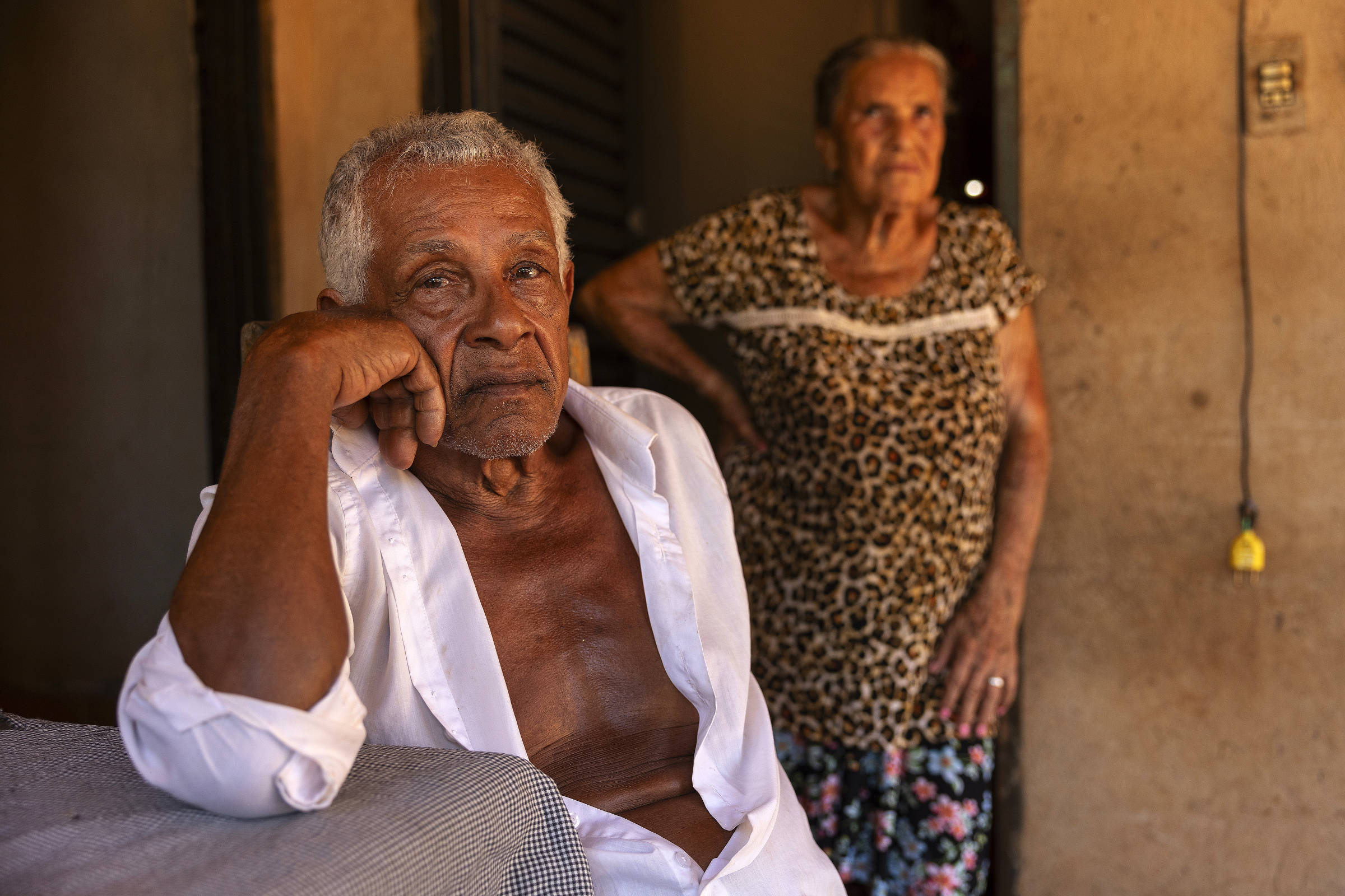 Retrato de homem negro, grisalho, sendo à meda e usando camisa branca aberta. Ao fundo, mulher grisalha usando blusa com estampa de pele de onça