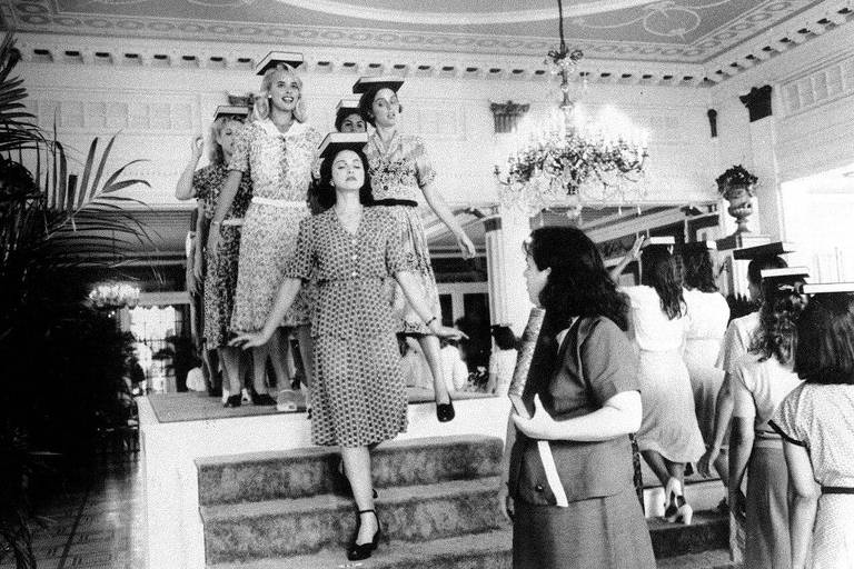 Foto em preto e branco mostra várias mulheres com vestidos dos anos 1940 caminhando com livros equilibrados sobre a cabeça. Bem à frente está Madonna, descendo degraus de uma pequena escada