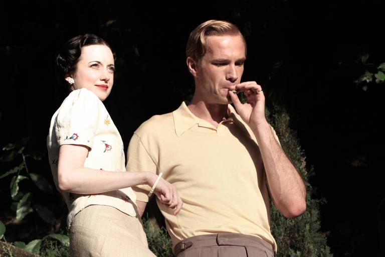 Um casal bem vestido com roupas dos anos 1940 desce por uma trilha em meio a uma mata