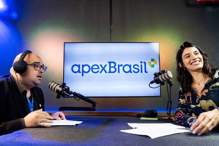 Homem está falando em microfone e mulher olha para a frente sorrindo, ao fundo está uma televisão com o logotipo da ApexBrasil escrito