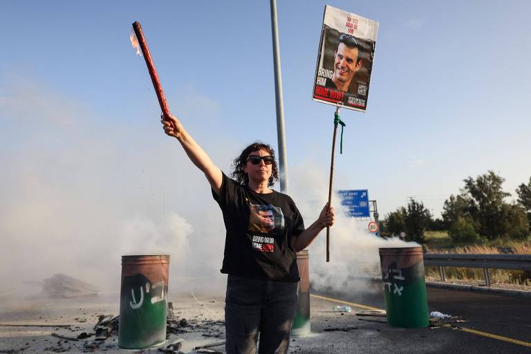 Mulher branca veste calça e camiseta preta, com a imagem de um refém israelense; com a mão direita ela segura uma tocha e com a outra um cartaz com a foto do homem sequestrado