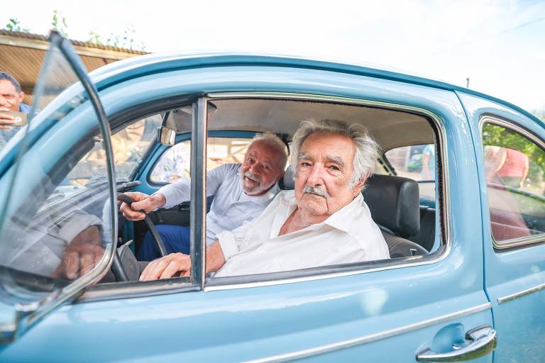 A imagem mostra dois homens dentro de um carro antigo azul. O homem à esquerda está dirigindo e sorri, enquanto o homem à direita, com cabelo grisalho, observa. O carro tem janelas abertas e o ambiente ao fundo é iluminado, sugerindo um dia ensolarado.