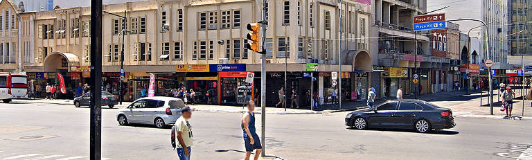 Cruzamento de avenidas, com dois pedestres no centro da imagem; prédio histórico de pintura bege ao fundo