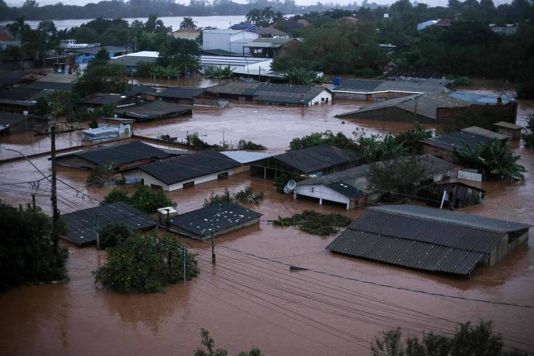 A imagem mostra uma área residencial gravemente afetada por uma enchente do rio Jacuí, em Eldorado do Sul (RS), com as águas turvas submergindo parcialmente as casas e a vegetação ao redor