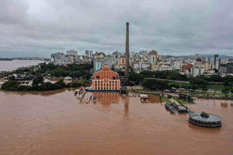 A imagem captura a cidade de Porto Alegre e o lago Guaíba, destacando uma construção histórica de tijolos aparentes à beira da água. O céu nublado e as águas barrentas do rio complementam o cenário urbano ao fundo, com edifícios modernos que se erguem por trás da chaminé industrial solitária.