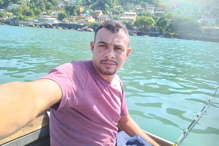 Carlos tira foto selfie em barco, ao fundo está o mar e uma ilha com casas e vegetação tropical