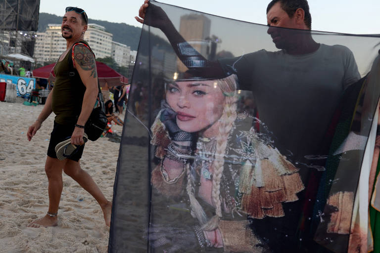 Homem segura tecido transparente com a foto da cantora Madonna na praia de Copacabana. Movimentação de público ao fundo.