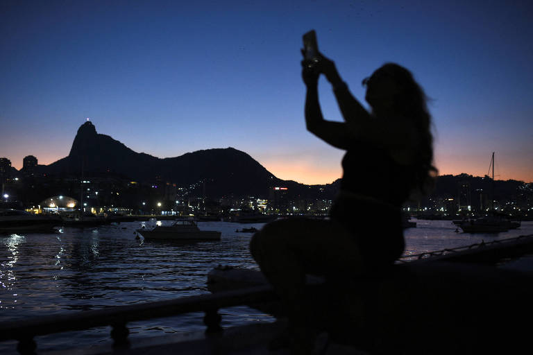 Silhueta de uma pessoa é vista segurando um celular durante op fim de tarde; paisagem mostra o mar com movimento de barcos e o morro ao fundo.