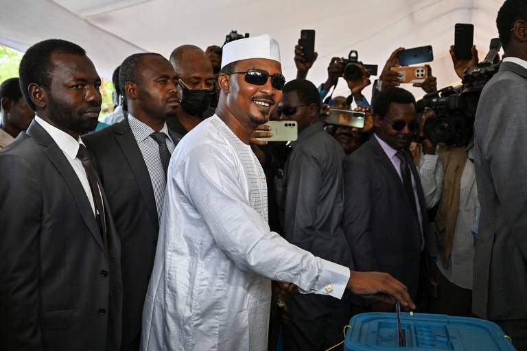 O candidato às eleições presidenciais de Chade, Mahamat Idriss Deby Itno, um homem negro vestido com roupa branca, rodeado por outros homens de terno, deposita seu voto em urna eleitoral


