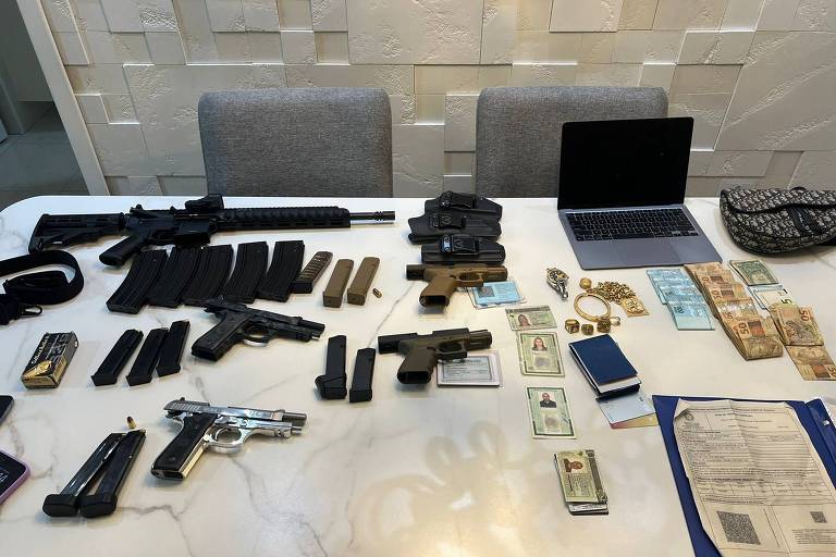Em uma mesa branca, armas, munições, notas de dinheiro, computadores e documentos são enfileirados