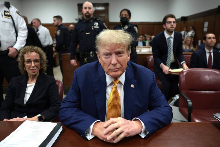 Donald Trump, um homem branco de cabelo branco usando terno, está sereno sentado em uma mesa de madeira, com as mãos cruzadas à frente; ao seu lado direito há uma mulher sentada, e ao fundo, alguns homens, entre eles um policial