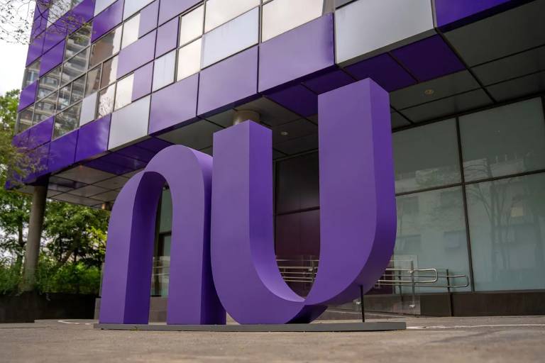 A imagem mostra um grande logotipo roxo em formato de "N" e "U" em frente a um edifício moderno com fachada de vidro