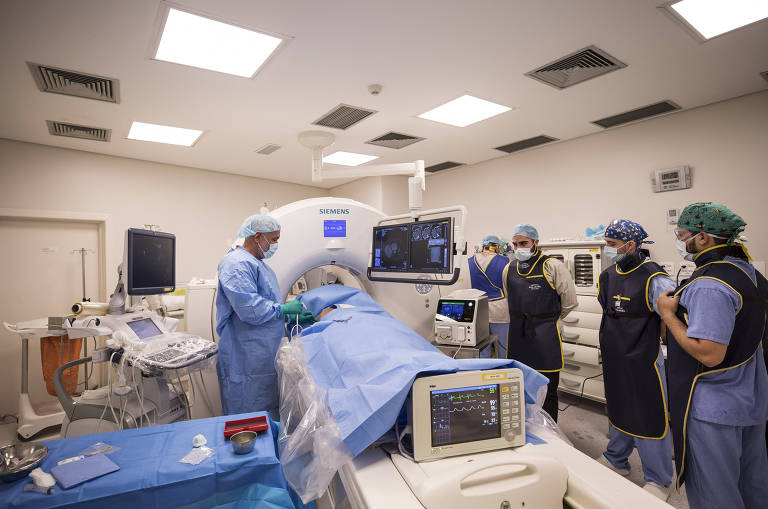 Imagem mostra médico de um lado do paciente e outras três pessoas da equipe, todos paramentados, durante um procedimento.