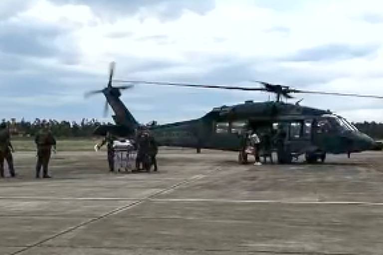 Helicóptero no chão com pessoas à frente. A aeronave está mais ou menos no meio da foto, com chão de cimento em primeiro plano e céu com nuves ao fundo