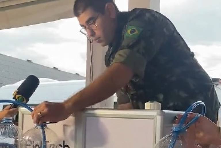 Militar das Forças Armadas usa farda verde e óculos, ele manuseia purificadores de água brancos e segura uma mangueira azul. Na parte de trás do equipamento há uma bandeira do Brasil.