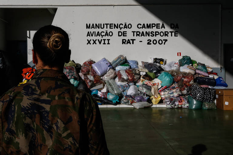Mulher soldada usa farda do Exército Brasileiro e aparece de costas, com cabelo preso. Ao fundo, sacolas estão empilhadas e dentro delas há objetos para doação.