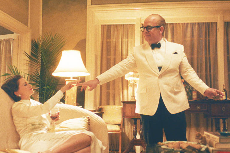 Imagem de um episódio da série. À esquerda, Naomi Watts, como Babe Paley, está jogada em um sofá numa sala de estar de um apartamento grande e chique, dos anos 1960. Ao seu lado, em pé, Tom Hollander como Truman Capote, vestindo um smoking branco, lhe entrega uma taça