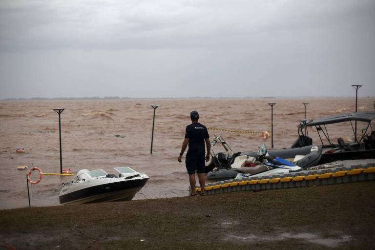 Homem, de costas para a câmera, observa as águas barrentas do lago Guaíba cercado por embarcações que atuam nos resgates