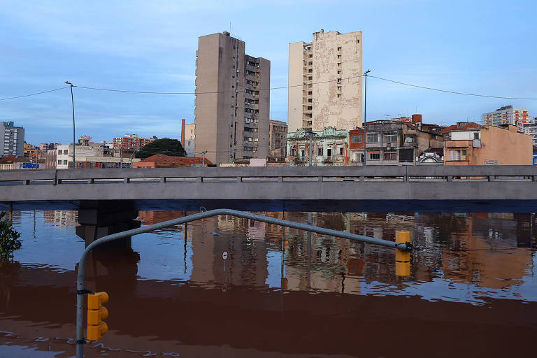 Uma ponte cruza a foto de lado a lado. Abaixo, um espelho d'água marrom das cheias. Acima dois prédios grandes e outros menores.