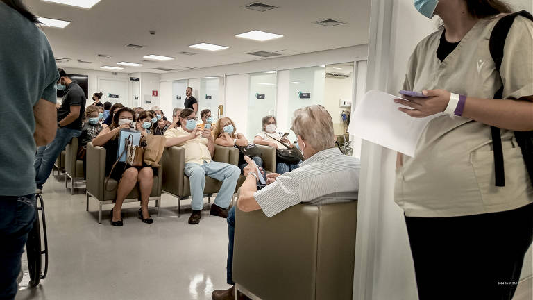 Pessoas com máscara sentadas e outras em pé aguardam em sala cheia de pacientes