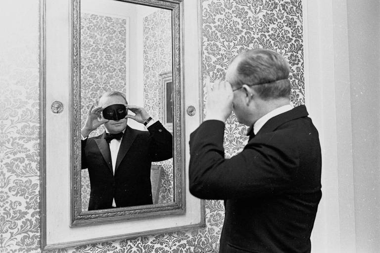 Um homem se encara no espelho para experimentar uma máscara negra sobre os olhos; ele é branco, tem os cabelos curtos penteados para traz e e usa um smoking
