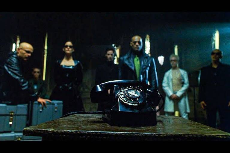 À frente, um telefone preto em uma mesa; atrás, os personagens do filme