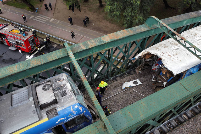 Uma vista aérea mostra os dois trens encima do viaduto. Ao meio, um trabalhador de capacete amarelo e um de capacete vermelho. O trem da direita está parcialmente despedaçado. 