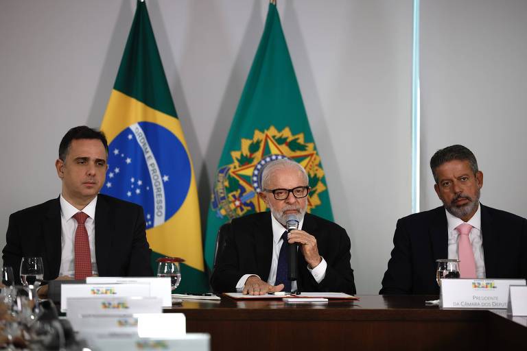 Três homens sentados atrás de mesa de madeira. Eles vestem terno escuro, camisa branca e gravata. Atrás deles, a bandeira do Brasil e símbolo do Brasão das Armas