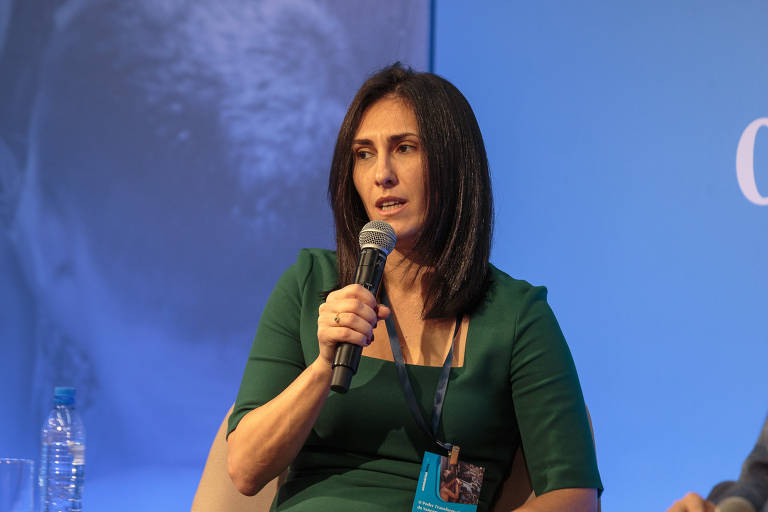 A imagem mostra uma mulher de cabelos escuros e lisos, vestindo uma blusa verde, segurando um microfone enquanto fala em um evento. O fundo é azul.