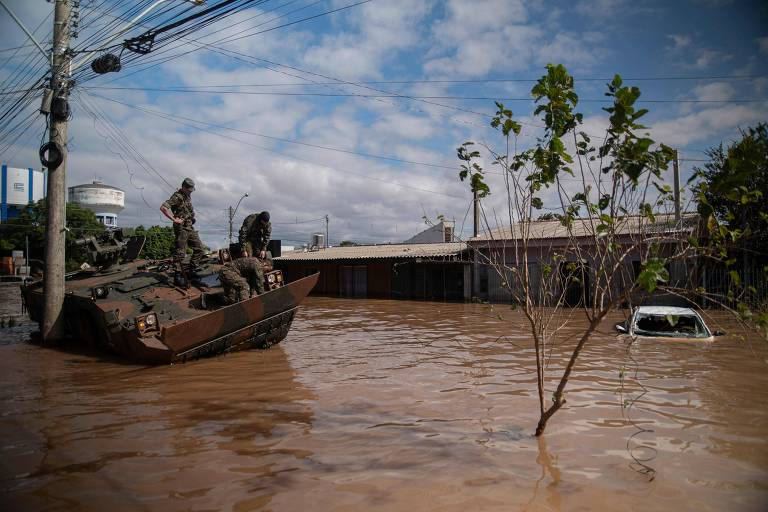 tanque do exército está em rua totalmente alagada, ao lado de carro que está afundado até o teto em água de cor lamacenta 