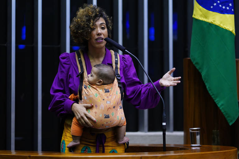 mulher negra com bebê em canguru laranja. ela usa camisa roxa e ao lado se vê a bandeira do brasil