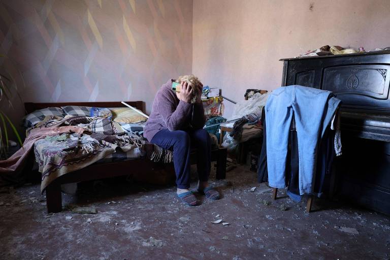Morador sentado na cama com a cabeça apoiada entre as mãos. o quarto está bagunçado pelo impacto do míssil na vila