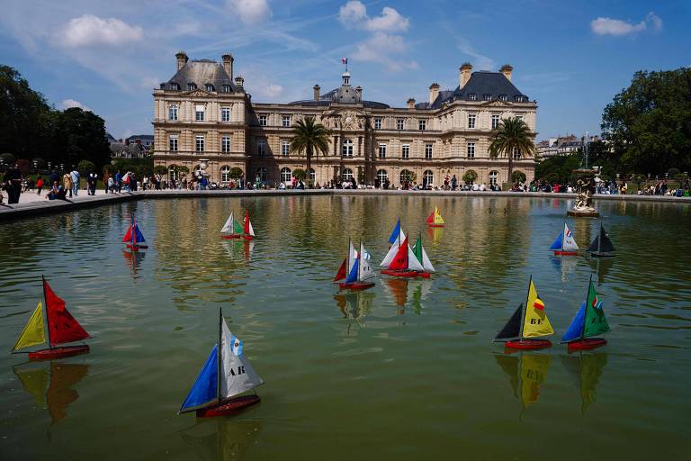 imagem geral mostra barcos à vela são vistos no Jardin du Luxembourg em Paris; edificação, árvores e céu azul ao fundo.