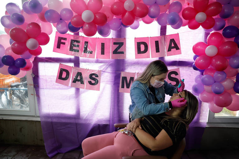Uma mulher, que está em pé e usando máscara, faz procedimento estético em outra que está sentada em uma cadeira reclinada em ambiente decorado com balões com os dizeres 'Feliz Dias das Mães'