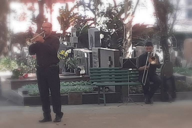 Em foto colorida, um duo formado por um trompete e um trombone toca em meio a sepulturas do cemitério do Redentor, no bairro do Sumaré, em São Paulo