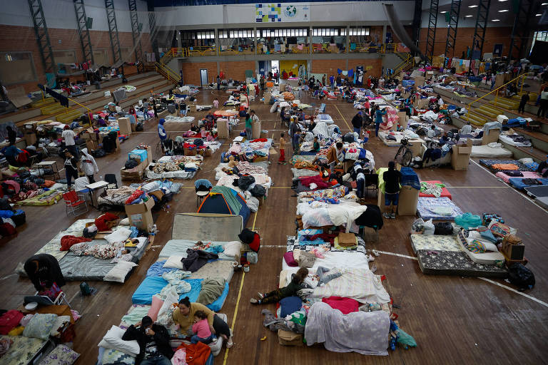 Na foto, dezenas de pessoas estão em um ginásio. No chão, colchões e roupas de cama