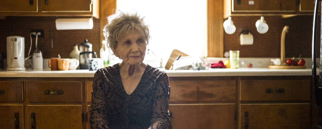 mulher idosa em cozinha com móveis de madeira cruza os braços enquanto olha obliquamente para o lado, com mãos pousadas sobre a mesa