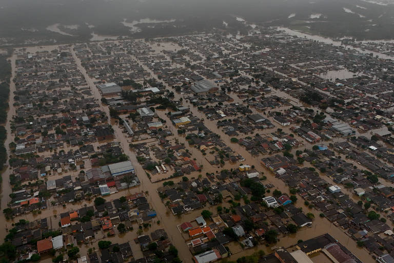 Imagem aérea de uma cidade afetada por inundações, mostrando ruas e casas submersas em água. O céu está nublado, e a água cobre grande parte da área urbana, com algumas edificações visíveis acima do nível da água.
