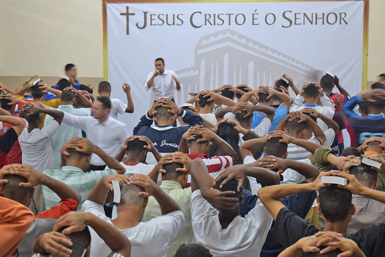 Homens aparecem posicionados em fileiras e com as duas mãos na cabeça. Um homem que usa camisa branca fala no microfone e, ao fundo, há um quadro branco com a frase: Jesus Cristo é o Senhor.