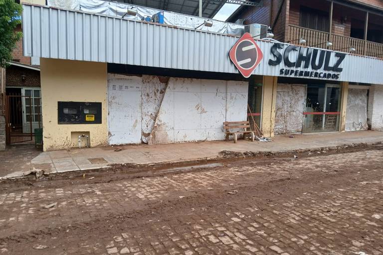 Imagem mostra a fachada do supermercado Schulz, em Sinimbu, com madeiras fechando as portas levadas pela enchente e a rua ainda com uma camada de barro